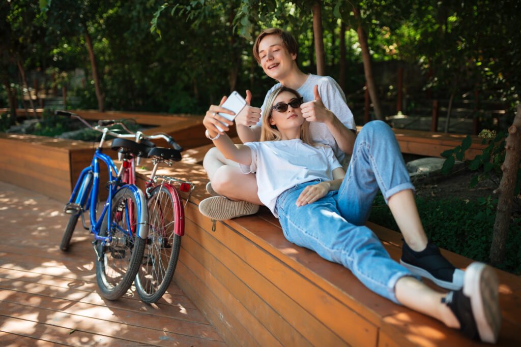 Lifestyle young joyful couple spending time in park with bicycles nearby. smiling boy sitting on bench in park with pretty girl with blond hair that leaning on him and taking selfie together